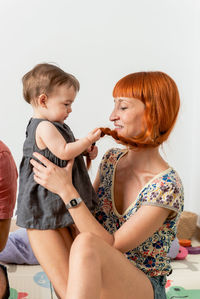 Cheerful young mother with long red hair in casual clothes smiling and embracing adorable baby while sitting on floor near sofa at home