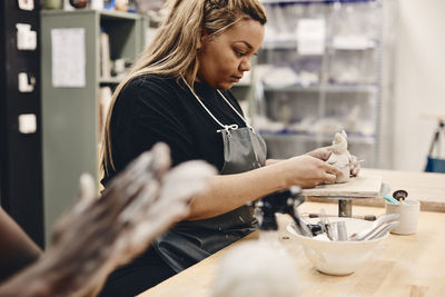 Young woman with work tool molding clay sitting at table in art class