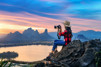 Rear view of woman standing on rock by lake against sky during sunset