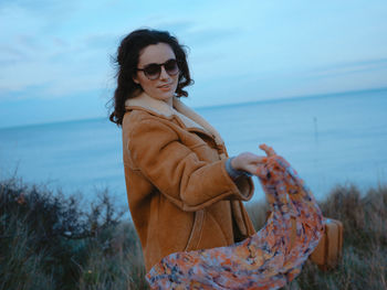 Young woman wearing sunglasses standing at beach against sky