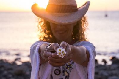 Smiling woman wearing hat giving flower against sea