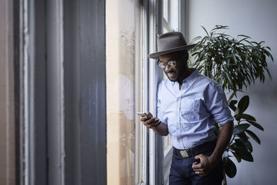 Smiling businessman using mobile phone while standing by window in creative office