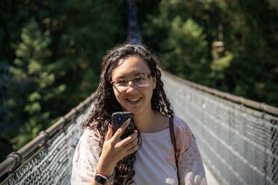 Portrait of young woman standing against trees with cell phone