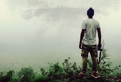 Rear view of man with camera standing on mountain in foggy weather