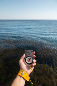 A man's hand with a wristwatch bracelet holds a magnetic compass against the background of the sea