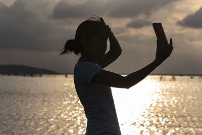 Silhouette woman taking selfie by sea against cloudy sky during sunset