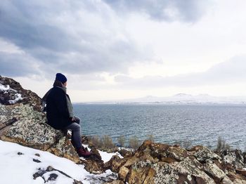 Side view of man sitting on rocks at beach against sky