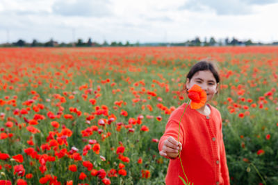 Dark haired girl standing in the poppy field holding out the flower