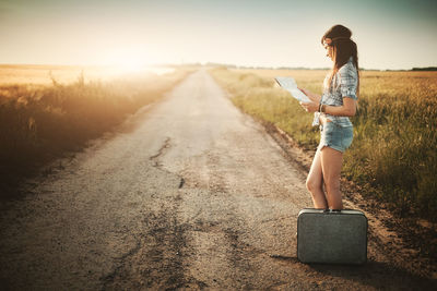 Side view of woman reading map standing by suitcase on road