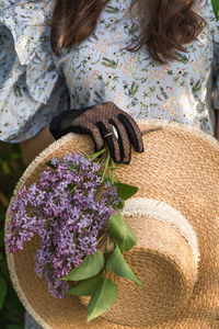 Woman's hand in vintage lace glove holds a straw hat with branch of lilac flowers. cottagecore.