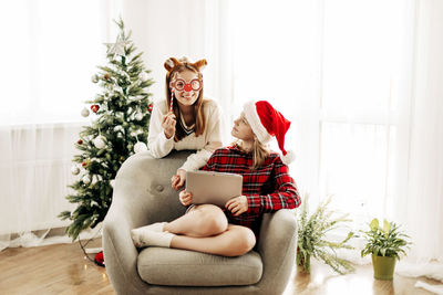 Two joyful sisters are sitting at home near the christmas tree and congratulating their friends 