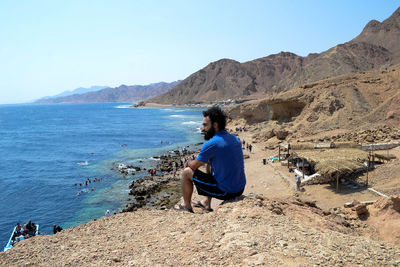 Man sitting on rock looking at mountains against clear sky