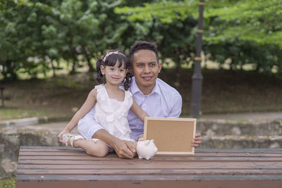 Smiling father and daughter holding piggy bank while sitting outdoors