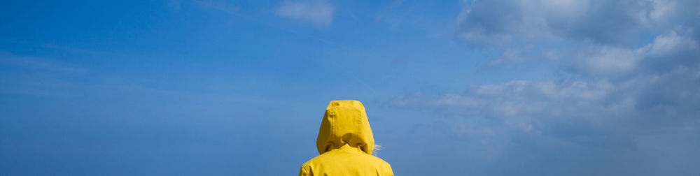 Rear view of woman on a sandy beach wearing a yellow hooded jacket and facing ocean withblue sky.