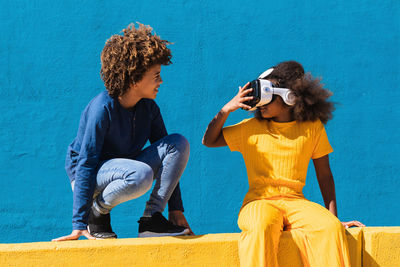 Low angle full body of african american boy communicating with girlfriend wearing vr headset while playing virtual game near colorful wall