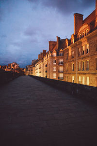 Walkway by illuminated buildings against cloudy sky at dusk