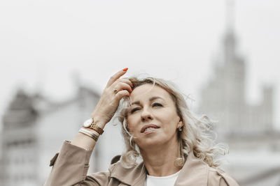 A middle-aged blonde woman with curly hair stands on the river embankment in the city.