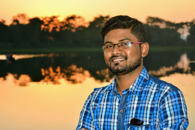 Portrait of young man standing against lake during sunset