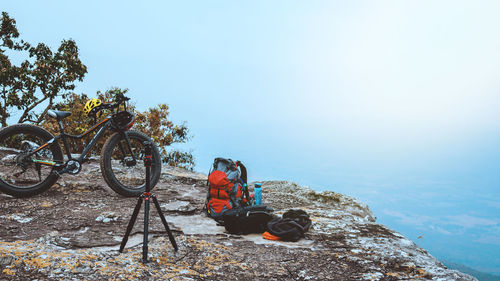 Bicycle by backpack with tripod on mountain against sky