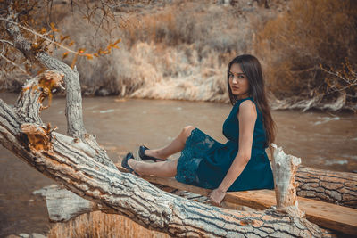 Portrait of beautiful young woman sitting on wood by fallen trees at river