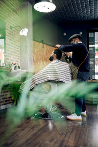 Side view full body of bearded barber spraying hair of male customer sitting on chair in modern barbershop during procedure