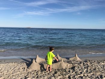 Rear view of boy standing by sandcastle on shore beach against sky