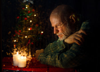 Close-up of thoughtful senior man sitting by illuminated christmas tree at home