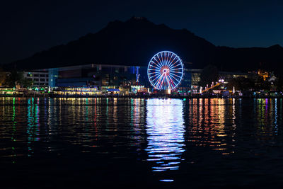 Illuminated ferris wheel by lake against sky at night