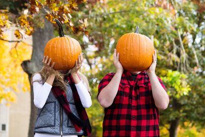 Man and woman hiding face with pumpkins against trees in yard