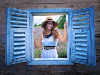 Woman wearing white dress and sun hat looking out through a wooden window