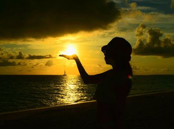 Optical illusion of silhouette woman holding sun at beach against sky during sunset