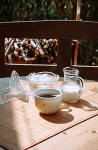 A cup of coffee stands outside on a table next to a jug of milk and a sugar bowl with lump sugar