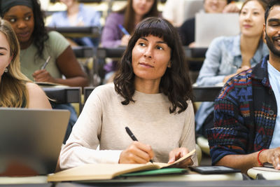Female student writing in book while sitting amidst friends at lecture hall