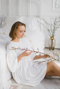 Young woman in a white dress holds an easter branch in her hands and sits at home by the bed