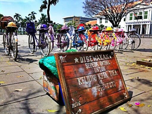 text, western script, communication, building exterior, built structure, non-western script, information sign, sunlight, street, day, architecture, outdoors, tree, sign, transportation, wood - material, capital letter, incidental people, bicycle, land vehicle