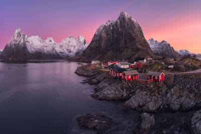 From above breathtaking view of hamnoy village with red and white houses located on coast of calm sea against snowy mountain ridge and sunset sky on lofoten islands, norway