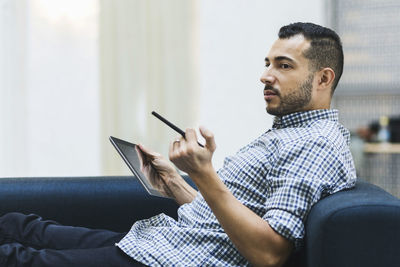 Side view of businessman holding digital tablet while relaxing on sofa at office