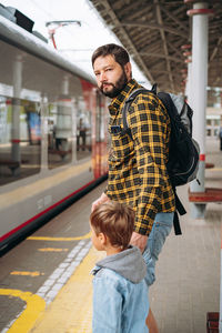 Caucasian man with backpack holding little son by hand standing at platform going to take train.