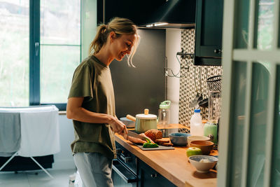 A young happy blond smiling woman cooking food in a home kitchen.