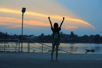 Silhouette woman with arms raised standing at riverbank against sky during sunset