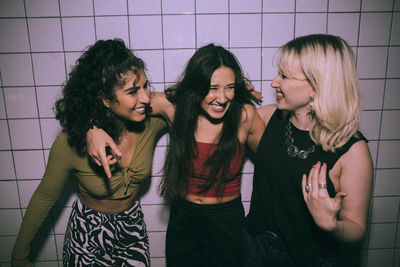 Cheerful young multiracial female friends standing with arms around against white wall at nightclub