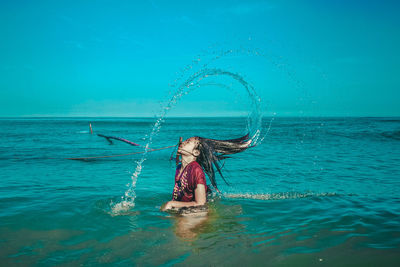 Side view of woman tossing hair in sea against blue sky