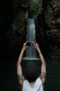 Rear view of man photographing waterfall with mobile phone