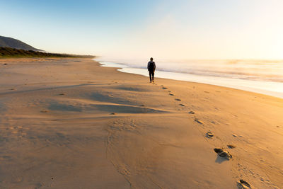 Rear view of man walking at beach against sky during sunset