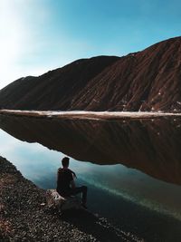 Man sitting at lakeshore by mountains against sky