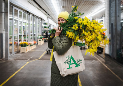 Positive woman in yellow cap and large bouquet of mimosa flowers at flower market
