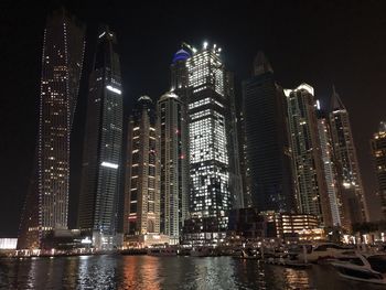 Illuminated modern buildings by sea against sky at night