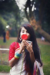 Portrait of young woman holding flower while standing against trees at park