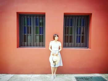 Woman holding hat while standing by orange wall with windows