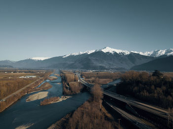 Scenic view of highway next to ricer against snowcapped mountains and against sky